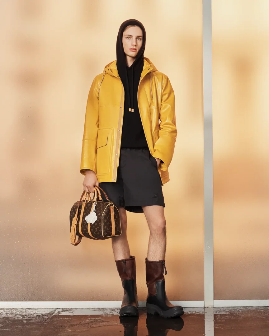 Genç bir erkek model, LOUIS VUITTON Ön Koleksiyonu İlkbahar-Yaz 2027 görünümünde, parlak sarı kapüşonlu deri bir ceket, siyah şort ve iki renkli uzun botlar giyerek, ikonik monogram desenli bir seyahat çantasını tutarken, ıslak görünümlü bir zeminde poz veriyor.