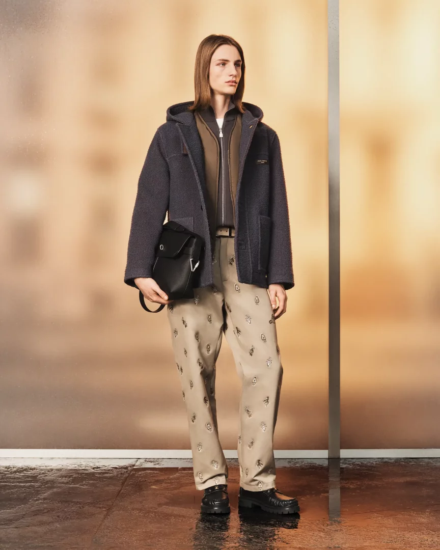 Louis Vuitton İlkbahar-Yaz 2027 erkek koleksiyonundan bu görünümde, uzun saçlı genç bir model, dokulu koyu gri kapüşonlu bir ceket, kahverengi fermuarlı bir üst, tüm yüzeyinde metalik işlemeler olan bej bir pantolon, kalın tabanlı siyah makosenler ve sol kolunda siyah deri bir omuz çantasıyla şık bir kombin sergiliyor.