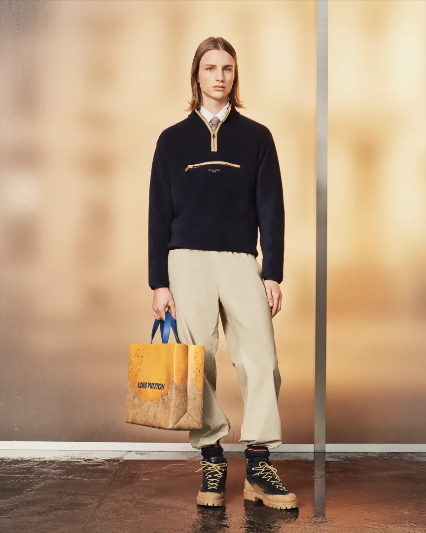 Uzun saçlı genç bir erkek model, Louis Vuitton pre-koleksiyonunun bir parçası olarak, beyaz gömlek ve kravat üzerine giydiği lacivert fermuarlı polar üst, bej bol pantolon ve kalın tabanlı botlarıyla dururken, kum ve toprak efektli Louis Vuitton logolu bir çantayı taşıyor.