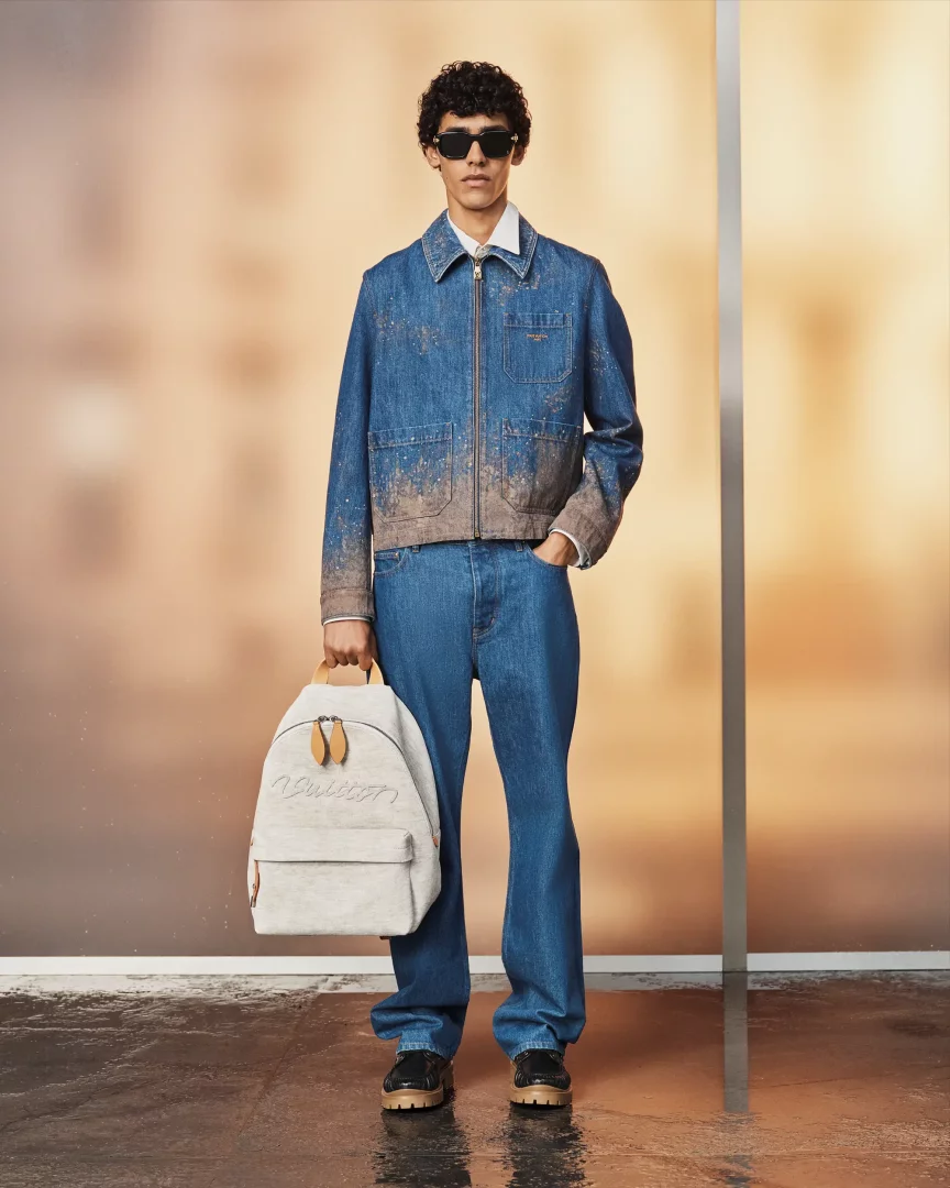 Siyah güneş gözlüklü, kıvırcık saçlı genç bir model, Louis Vuitton İlkbahar/Yaz 2027 ön koleksiyonundan topraklı efektli mavi denim ceketi, bol paça kot pantolonu ve kalın tabanlı ayakkabılarıyla poz verirken sağ elinde kahverengi deri detaylı bej bir sırt çantası tutuyor.