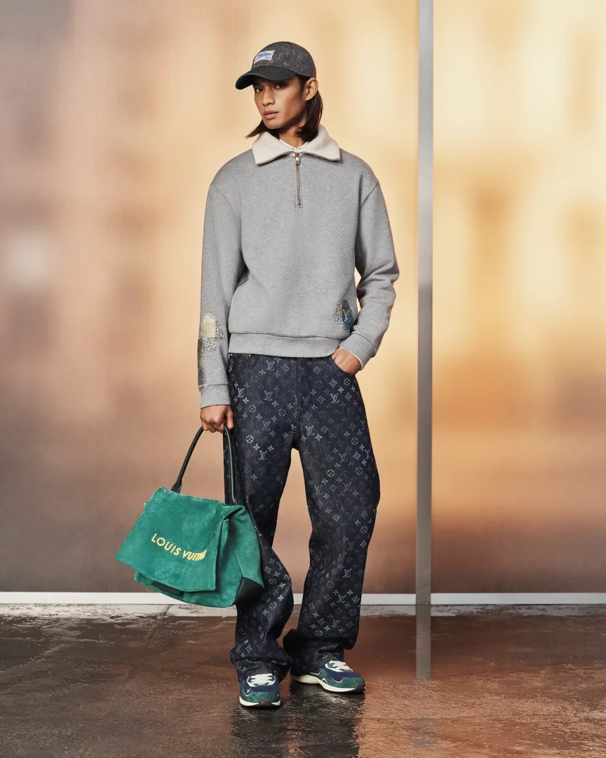 Model, Louis Vuitton İlkbahar-Yaz 2027 erkek ön koleksiyonundan bir kombini sergiliyor: açık renk yakalı gri yarım fermuarlı sweatshirt, manşetlerinde hafif işlemeler olan, Louis Vuitton monogram desenli koyu renk kot pantolon ve spor ayakkabılarla birlikte, altın renkli logosu belirgin zümrüt yeşili süet bir çanta taşıyor.