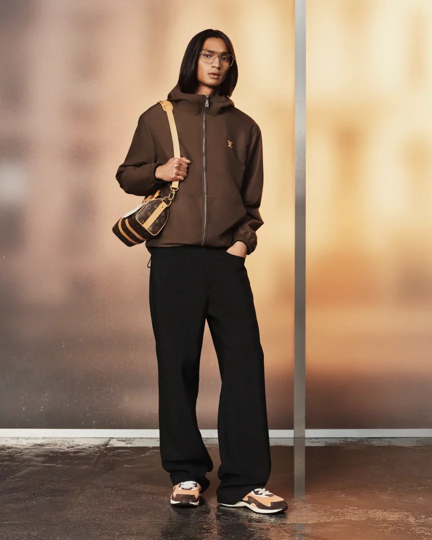 Louis Vuitton'un 2027 İlkbahar/Yaz erkek ön koleksiyonundan 26. görünümü sergileyen, kahverengi kapüşonlu fermuarlı ceket, siyah bol pantolon, spor ayakkabı ve LV monogramlı küçük omuz çantası taşıyan uzun saçlı bir model stüdyoda duruyor.