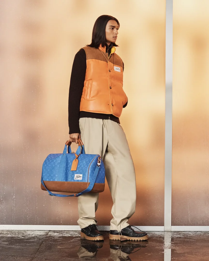 Louis Vuitton Erkek İlkbahar-Yaz 2027 Pre-Koleksiyonundan bir model, siyah kazak üzerine turuncu ve kahverengi şişme yelek, bej bol paça pantolon ve kalın siyah ayakkabılarla dikkat çekiyor; modelin elinde, zemine yansıyan ışıklarla parıldayan mavi renkli Louis Vuitton logolu seyahat çantası bulunuyor.