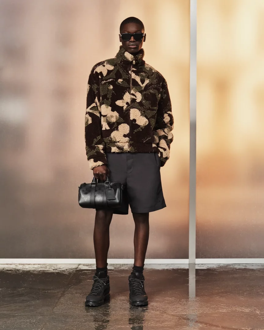 Louis Vuitton İlkbahar-Yaz 2027 Pre-Koleksiyonu'ndan bir model, koyu kahverengi, yeşil ve bej tonlarında kamuflaj desenli, fermuarlı kabarık bir ceket, koyu renk şort ve kalın tabanlı botlarla modern bir görünüm sergilerken, sağ elinde ikonik siyah Louis Vuitton çantasını tutuyor.
