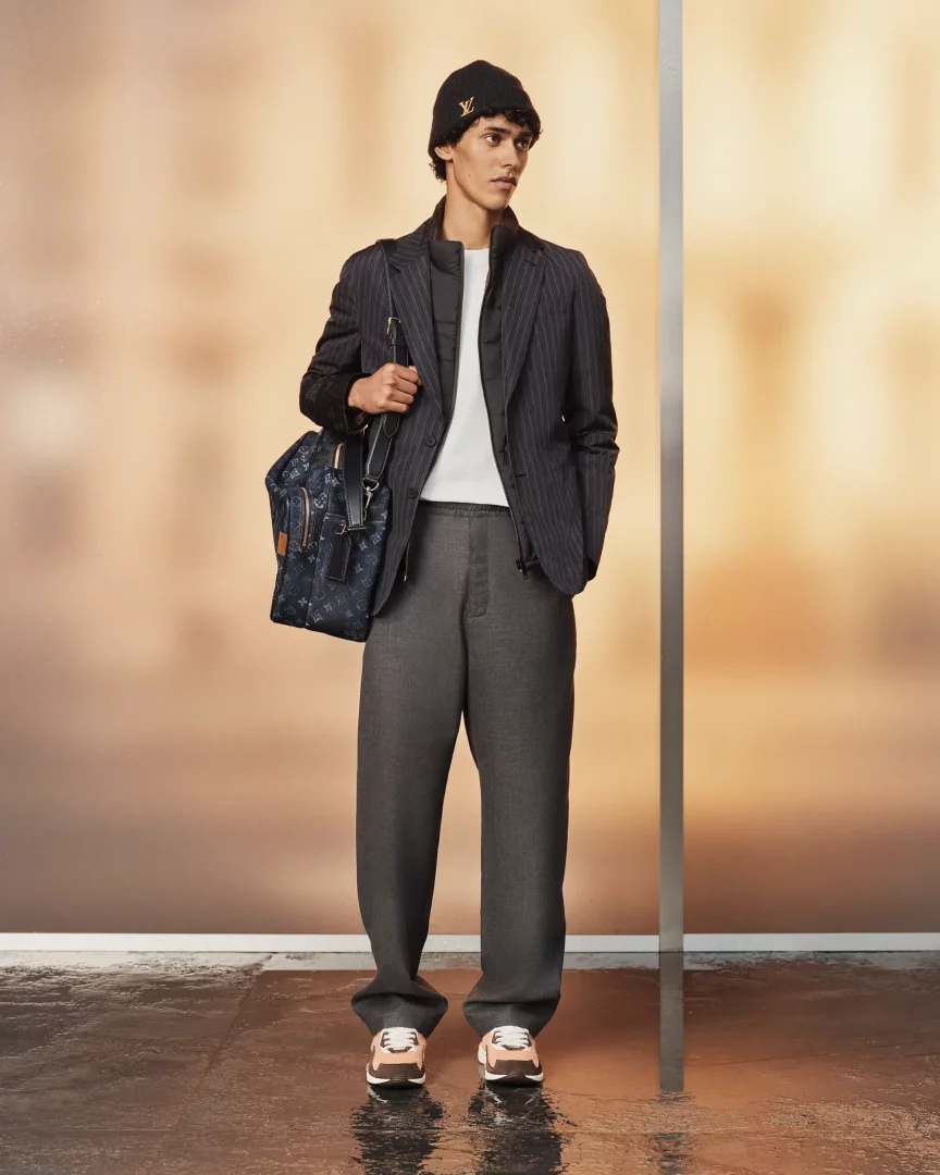 Louis Vuitton Pre-Koleksiyonu İlkbahar/Yaz 2027'den bir görünüm sergileyen genç model, siyah bere, çizgili blazer, bol gri pantolon, pembe-bej renkli spor ayakkabıları ve Louis Vuitton logolu lacivert çantasıyla, ışıklı bir arka plan önünde duruyor.