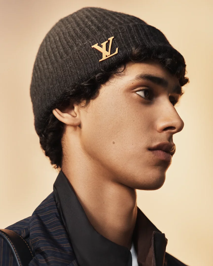 Louis Vuitton erkek koleksiyonundan, genç bir modelin profilinden çekilmiş yakın planda, başındaki fitilli koyu gri berede parlayan altın rengi LV logosu ve kıvırcık saçları öne çıkarken, zarif çizgili bir ceketle ilkbahar-yaz şıklığını sergiliyor.