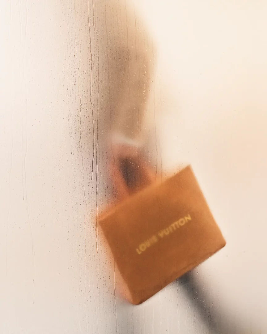 Buharlı ve ıslak camın arkasından flu bir şekilde görünen, üzerinde LOUIS VUITTON yazısı bulunan turuncu-kahverengi bir kutuyu tutan kişinin silüeti, Louis Vuitton Pre-Koleksiyonu Bahar/Yaz 2027 temasını yansıtan gizemli bir anı yakalıyor.