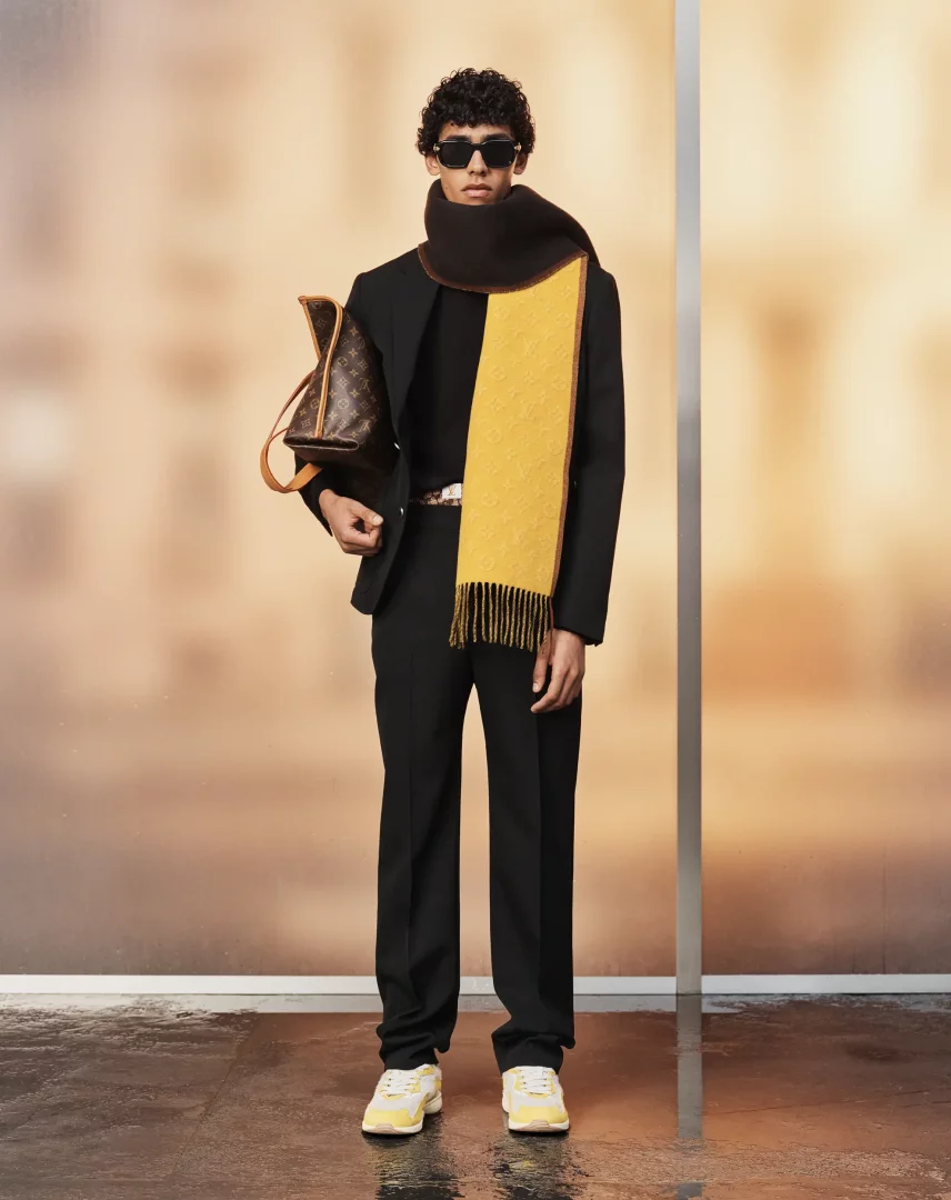 Louis Vuitton İlkbahar-Yaz 2027 erkek ön koleksiyonundan bir görünüm sergileyen kıvırcık saçlı, güneş gözlüklü bir model, siyah takım elbisesi ve siyah balıkçı yaka kazağı üzerine asimetrik sarı ve siyah renkli, logo desenli ve püsküllü gösterişli bir eşarp takmış, sol elinde Louis Vuitton monogramlı kahverengi bir çanta tutarken, sarı detaylı beyaz spor ayakkabıları ve açık renk kemeriyle yansımalı, sıcak tonlu bir arka plan önünde duruyor.