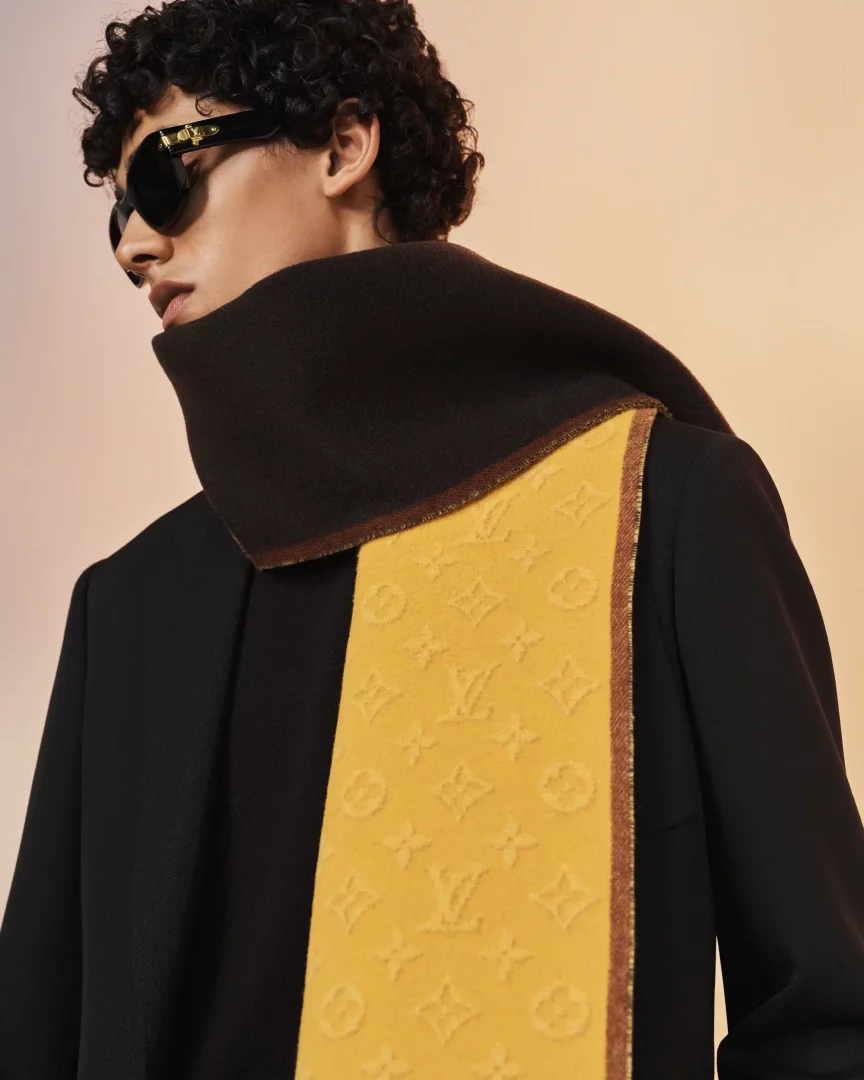 Louis Vuitton Erkek İlkbahar-Yaz ön koleksiyonundan, boynunda sarı ve kahverengi tonlarında, kabartma monogram detaylı bir Louis Vuitton şalı bulunan, siyah güneş gözlüklü ve kıvırcık saçlı bir modelin yakından çekilmiş zarif portresi.