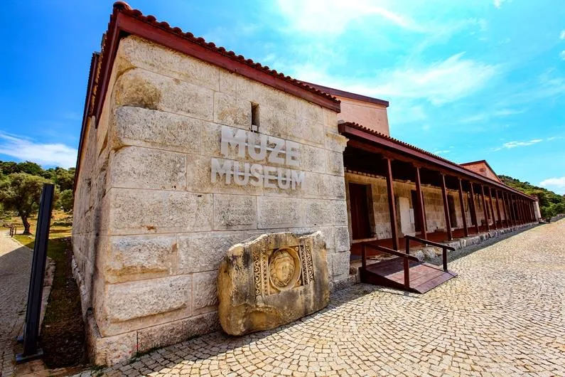 Parlak mavi gökyüzü altında, Likya Uygarlıkları Müzesi tabelasının da yer aldığı, taş bloklardan yapılmış binanın önünde kabartmalı antik bir taş eseri ve sağ tarafında uzun ahşap direkli bir revak ile parke taşlı bir yol uzanıyor.