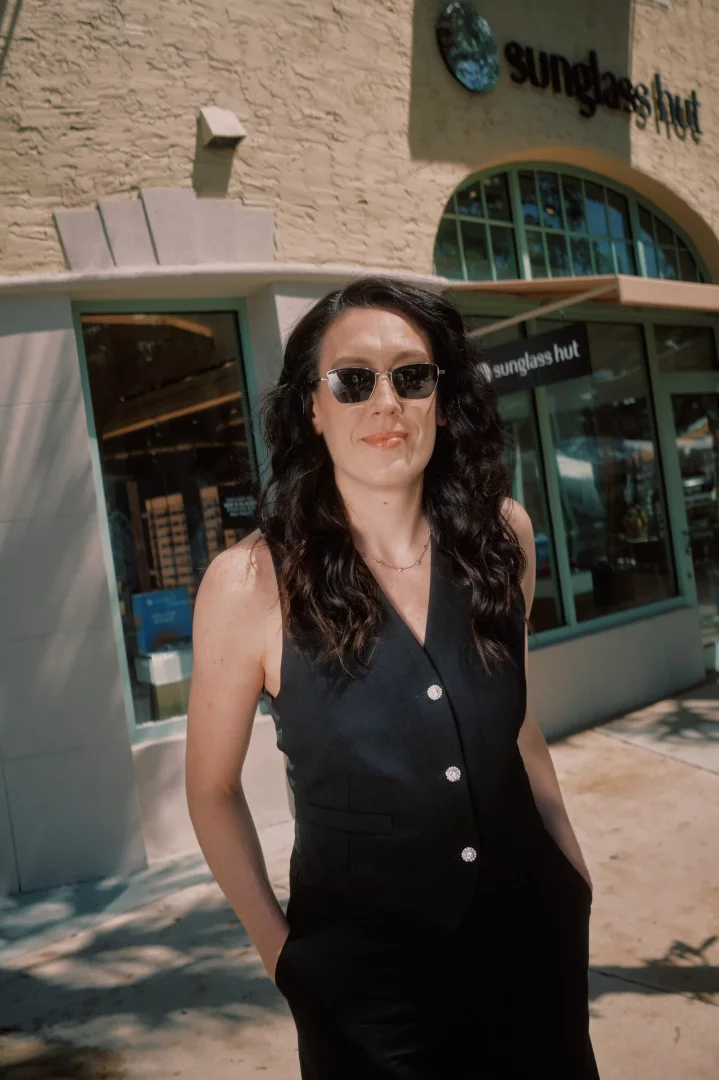 Güneşli bir günde, dalgalı saçlı ve siyah yelek giyen bir kadın, kendinden emin bir ifadeyle, üzerinde Sunglass Hut yazan mağazanın önünde durmuş, şık güneş gözlükleriyle objektife bakıyor.