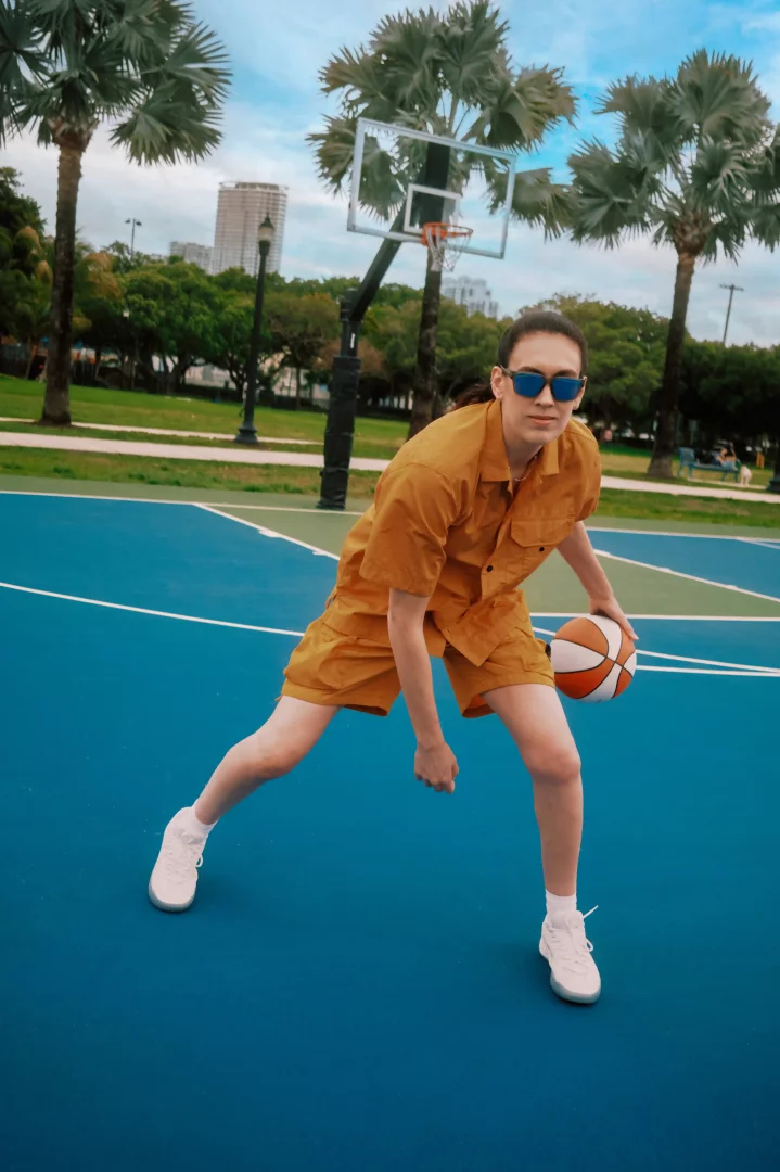 Mavi basketbol sahasında, sarı gömlek ve şort giymiş, güneş gözlüklü bir kişi dinamik bir duruşla basketbol topunu elinde tutuyor; arkasında palmiye ağaçları, basketbol potası ve şehir silüeti görünüyor.