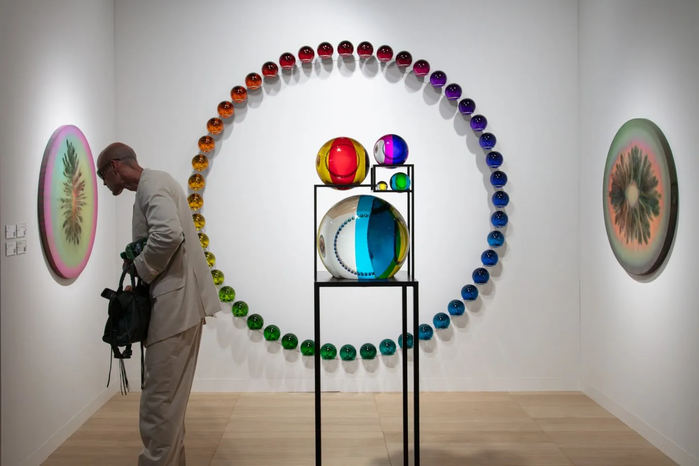 Beyaz duvarlı bir sanat galerisinde, açık renk takım elbiseli ve gözlüklü bir adam sol duvardaki pembe-yeşil tonlarda oval bir eseri yakından incelerken, merkezde siyah bir stand üzerinde parlak renkli cam küreler ve arka duvarda bir gökkuşağı renk geçişiyle düzenlenmiş sayısız küçük küreden oluşan çağdaş sanat eserleri dikkat çekiyor.
