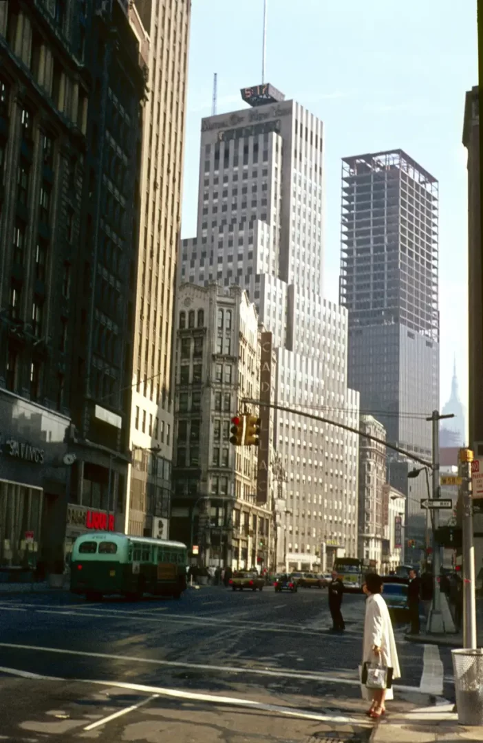New York şehrinin kalabalık bir caddesindeki tarihi atmosferi yansıtan bu geniş açılı görselde, hafif bir palto giyen bir kadın yaya geçidinde beklerken, yeşil bir otobüs ve sarı taksiler hareketli trafiği oluşturuyor; arka planda, National City Bank of New York tabelalı yüksek binalar ve inşa halindeki iskelet yapılar, 5:17'yi gösteren dijital saatle şehrin dinamik dokusunu sergiliyor.
