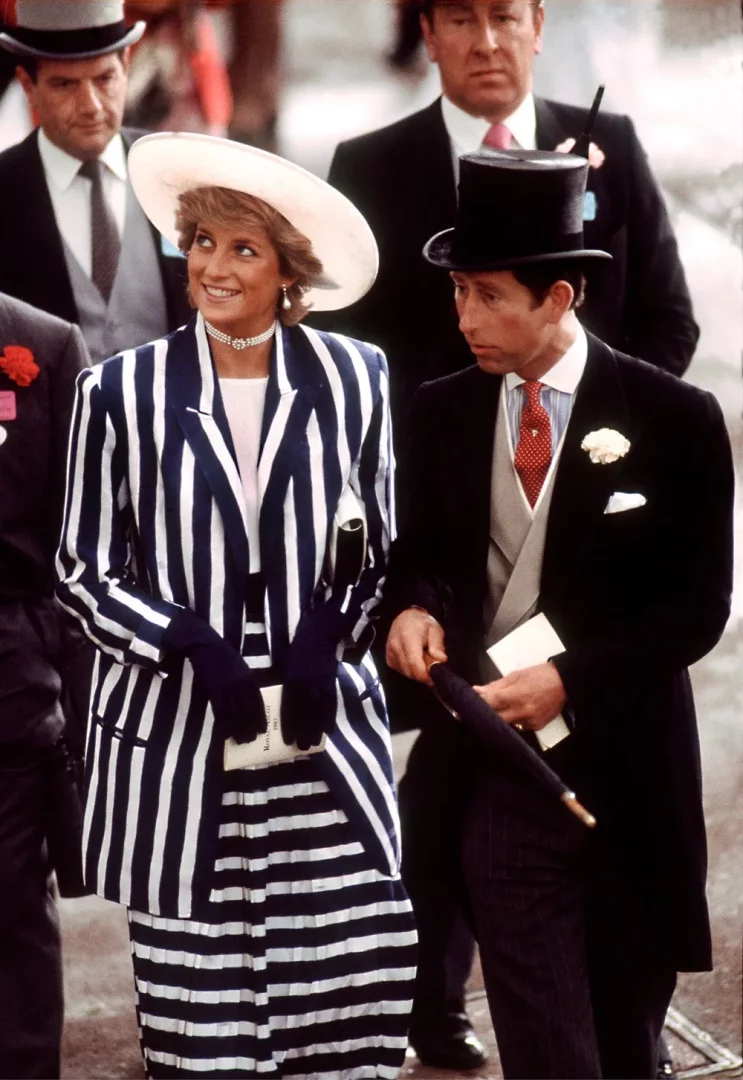 Prens Charles ve Prenses Diana, Royal Ascot etkinliğinde çekilen bu fotoğrafta, Prenses Diana beyaz geniş kenarlı şapkası, inci kolye ve küpeleriyle lacivert beyaz dikey çizgili ceketi ve yatay çizgili eteğiyle neşeli bir şekilde gülümserken, Prens Charles ise klasik siyah silindir şapkası, frakı ve kırmızı puantiyeli kravatıyla şemsiyesini tutarak yan tarafına bakıyor.