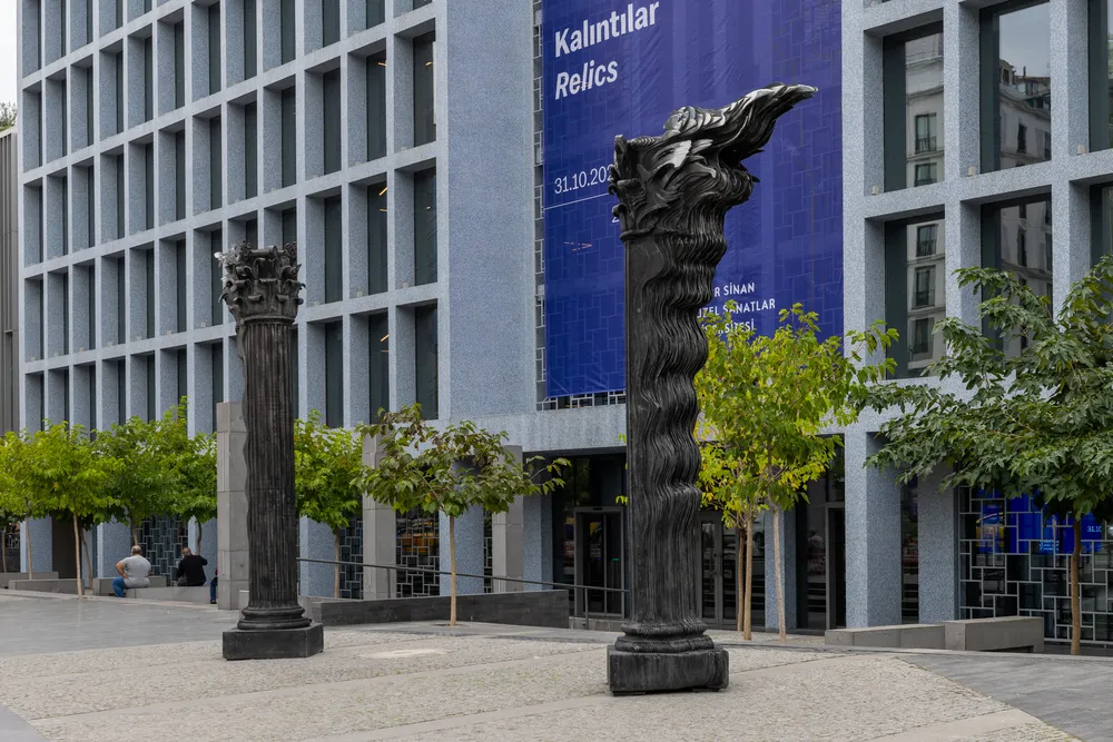 Gri kare pencereli modern bir binanın önünde, sol tarafta klasik Korint başlığına sahip, sağ tarafta ise alev veya akan saç gibi dalgalı soyut bir forma bürünmüş iki koyu renkli heykel sütun, Kalıntılar adlı açık hava enstalasyonunun dikkat çekici parçaları olarak, çevredeki genç ağaçlar ve banklarda oturan insanlarla birlikte bir şehir meydanında sergileniyor.