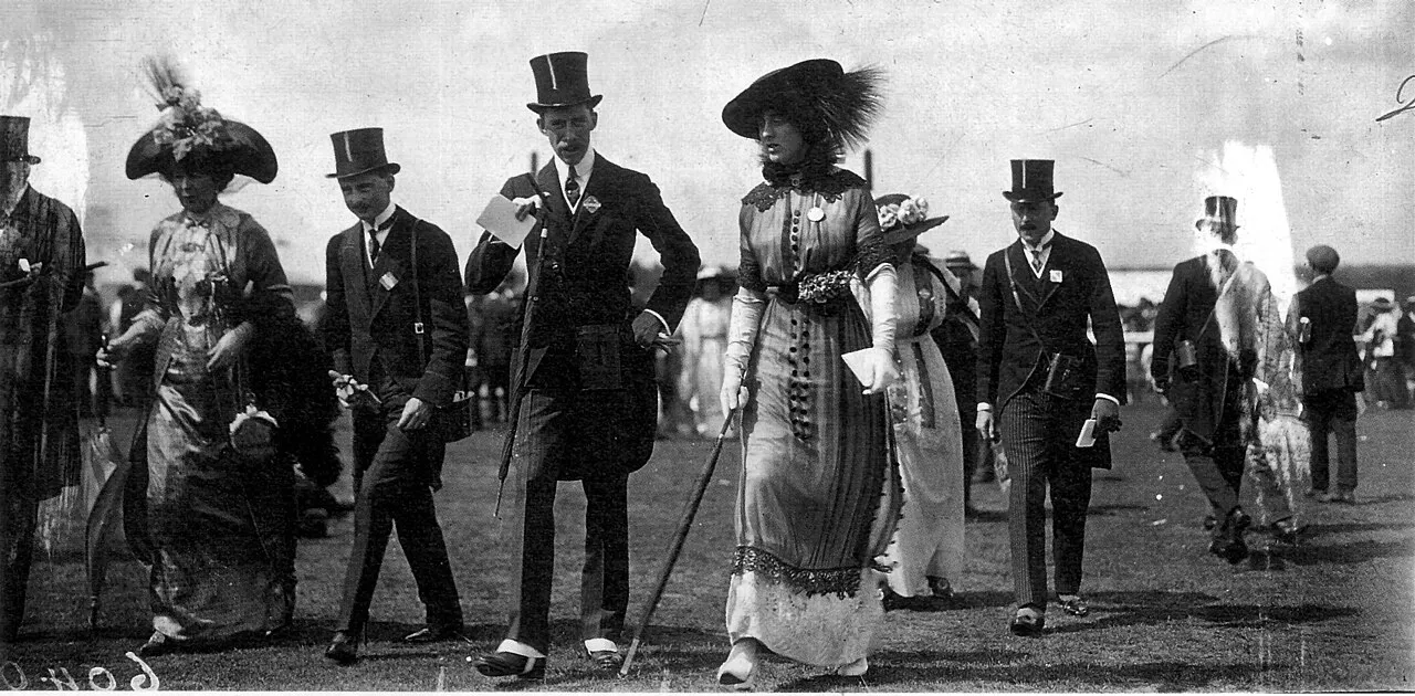 Geniş kenarlı tüylü şapkası ve uzun, süslemeli elbisesiyle Vita Sackville-West, 1912 Royal Ascot etkinliğinde silindir şapkalı ve fraklı beyler eşliğinde çim alanda yürürken, dönemin şıklığını gözler önüne seren tarihi bir fotoğraf.