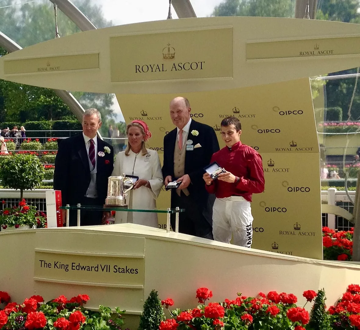 Royal Ascot'ta King Edward VII Koşusu ödül töreninde, takım elbiseli antrenör John Gosden'in de aralarında bulunduğu üç erkek ve beyaz giysili bir kadın, kupa ve madalyalarla poz verirken, arkalarında Royal Ascot ve sponsor QIPCO logoları belirgin şekilde görülüyor.