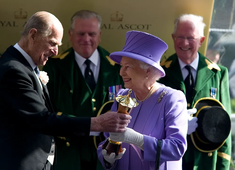 Royal Ascot etkinliğinde Prens Philip, mor elbisesi ve şapkasıyla zarif görünen Kraliçe II. Elizabeth’e gülümseyerek bronz ve altın renkli bir kupa takdim ederken, ikilinin arkasında etkinliğin enerjisini yansıtan diğer görevliler de gülümsüyor.