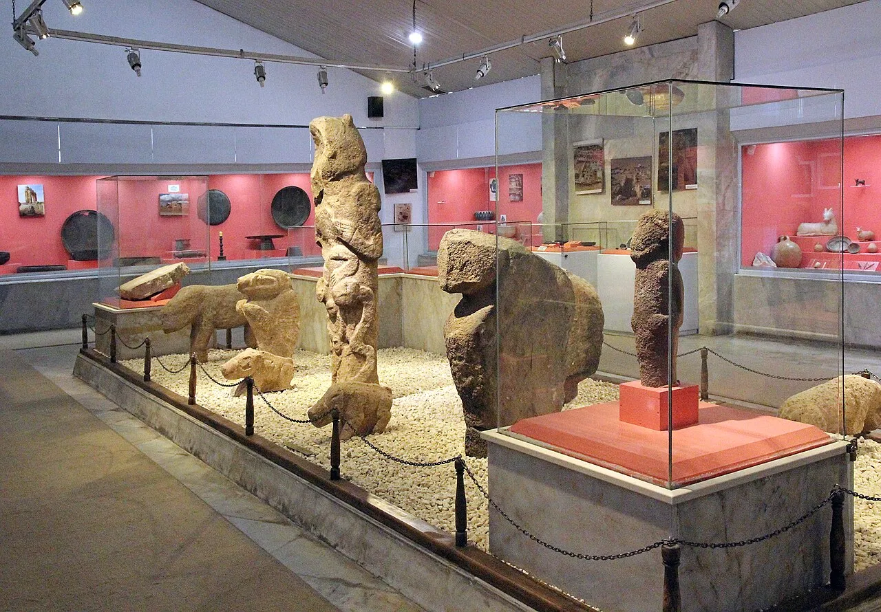 Şanlıurfa Arkeoloji Müzesi'nin geniş bir salonunda, çakıl taşlı zeminde zincirli bariyerlerle çevrili alanda özenle sergilenen, aralarında hayvan figürleri ve antromorfik dikilitaşların da bulunduğu çeşitli boyutlarda tarih öncesi taş heykeller, arka planda ise kırmızı panelli cam vitrinlerde küçük arkeolojik eserler görünmektedir.
