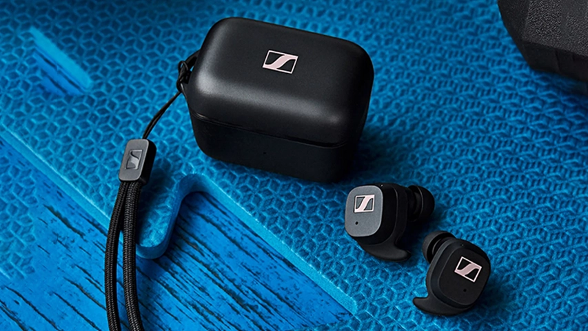 Mavi dokulu bir zemin üzerinde duran, siyah renkli Sennheiser Sport True Wireless kulaklıkların şarj kutusu, iki adet kulaklık ve kutuya takılı bir bilek kayışı, spor temalı bir ortamda ürünün kullanım alanına vurgu yapıyor.