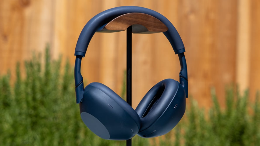 Ahşap destekli siyah bir standa asılı duran koyu mavi Sony WH-1000XM6 kulaklık, modern ve şık tasarımıyla öne çıkıyor; arka planda bulanık ahşap çit ve yemyeşil bitki örtüsü seçiliyor.