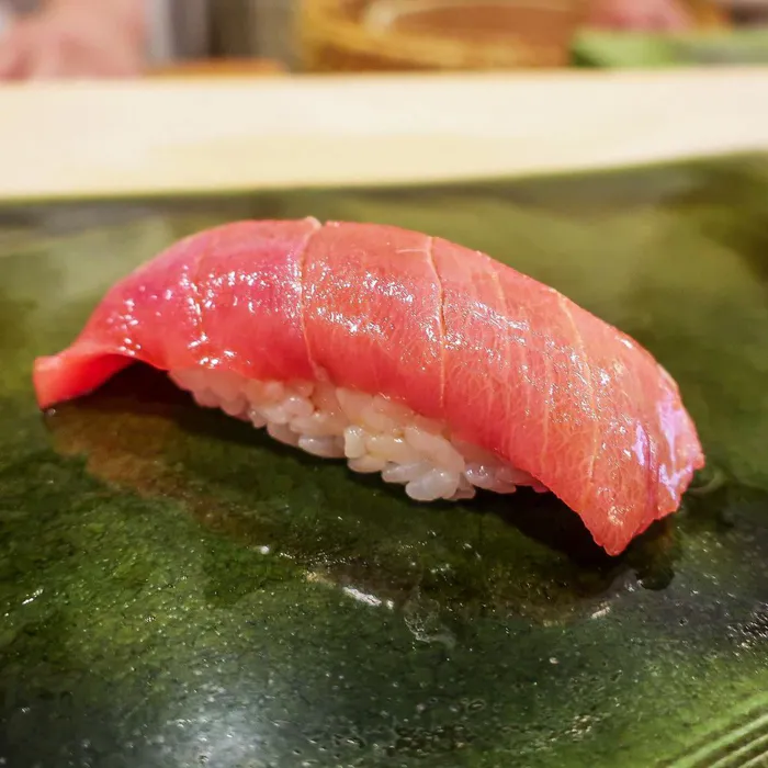 Mükemmel Sukiyabashi Jiro ustalığını yansıtan, koyu yeşil parlak bir tabakta sunulan, özenle hazırlanmış pirinç üzerine yerleştirilmiş, parlak kırmızı renkteki ton balığı dilimleriyle kaplı iştah açıcı bir nigiri suşi, arka planda hafifçe bulanık ahşap detaylar ile.