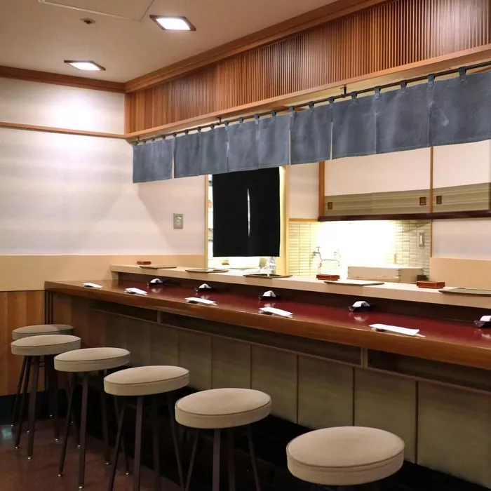 Sukiyabashi Jiro gibi ünlü bir suşi restoranının, zarif ahşap tezgahı boyunca düzenlenmiş altı minderli taburesi ve her biri için özenle yerleştirilmiş servis setleriyle geleneksel Japon zanaatkarlığını yansıtan iç mekanı.