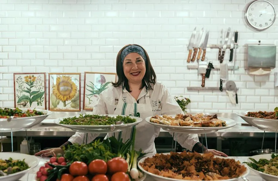 Beyaz karo duvarlı, profesyonel bir mutfak ortamında, gülen yüzlü bir şef kadın, önündeki ve elindeki taze salata, börek ve yöresel lezzetlerle dolu tabakları misafirlere sunmaya hazırlanıyor, adeta Beyrut'un Tawlet veya Souk el Tayeb ruhunu yansıtıyor.