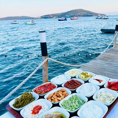 Kınalıada'daki Teos Restoran'da, ahşap bir iskelenin üzerinde sergilenen çeşit çeşit lezzetli meze tabağı, denizin üzerinde yüzen tekneler ve uzaktaki ada manzarası eşliğinde davetkar bir atmosfer sunuyor.