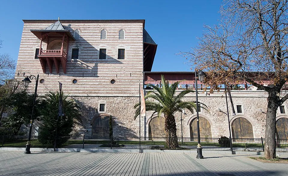 Güneşli bir günde çekilen bu geniş açılı görselde, üst katlarında yatay taş şeritleri ve solunda ahşap bir cumba ile öne çıkan Türk ve İslam Eserleri Müzesi'nin tarihi yapısı, alt katındaki kemerli ahşap kapıları ve önündeki palmiye ağaçları ile meydanın köşesinden tüm ihtişamıyla yükseliyor.