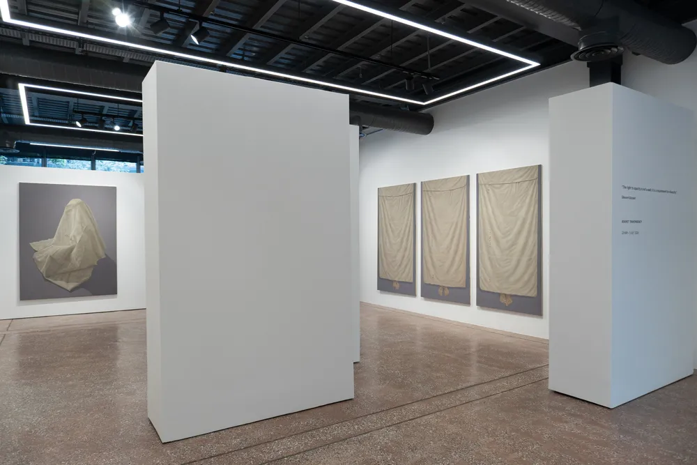 Şeffaflığa Karşı sergisi kapsamında, modern bir sanat galerisinde, beyaz bir duvarda gri zemin üzerinde örtülü bir figürü gösteren bir resmin ve ilerideki duvarda üç adet bej kumaşla kaplı dikey eserin sergilendiği, ayrıca sağdaki beyaz duvarda sergi başlığı ile tarihlerinin belirtildiği geniş bir alan.