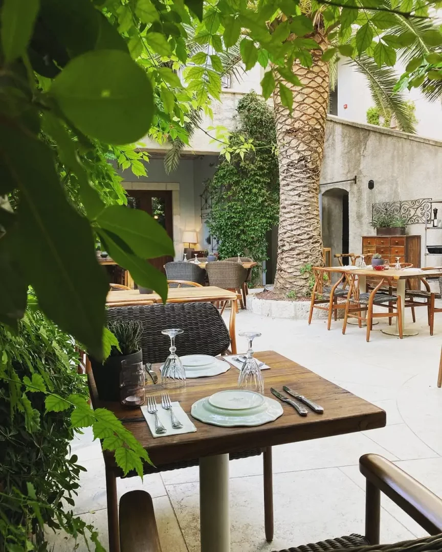 Alaçatı'nın en iyi mekanları arasında yer alan grilia restoranının yemyeşil bitkilerle çevrili, ortasında kalın gövdeli bir palmiye ağacının yükseldiği, şık ahşap masaların özenle servis için hazırlandığı huzurlu açık hava avlusu.