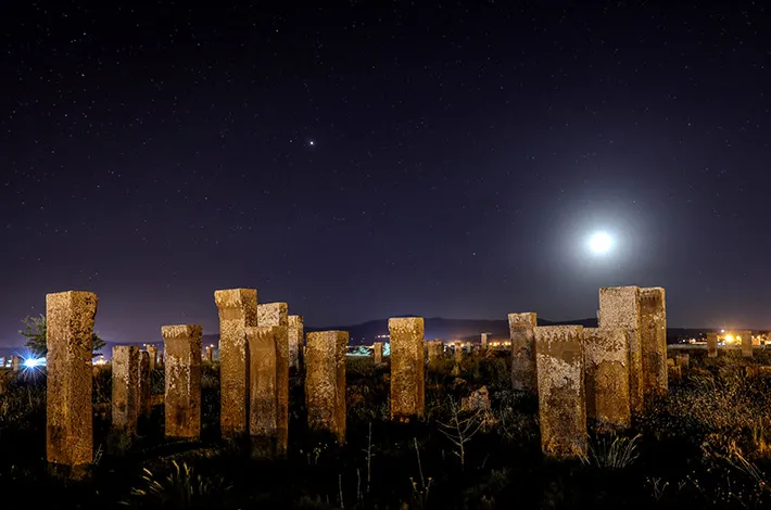 Karanlık bir gecede, Ahlat Selçuklu Mezarlığı'nda ay ışığının ve sayısız yıldızın aydınlattığı geniş bir alanda sıralanmış, yosunlu ve eski dikili taş mezar taşları, uzak şehir ışıklarıyla birlikte mistik bir atmosfer oluşturuyor.