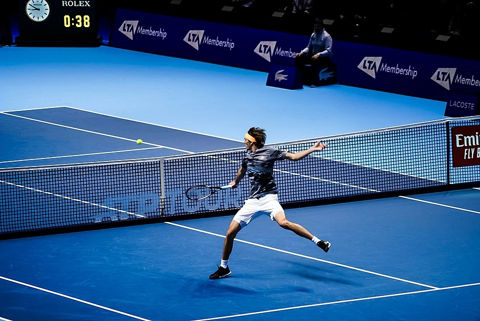 Mavi kortta hızlı bir topa vuruş yaparken enerjik bir duruş sergileyen tenisçi Alexander Zverev, tenis raketiyle fileye doğru uzanıyor; arkasında LTA Membership ve Lacoste gibi sponsor logoları ile Rolex skorboardu net bir şekilde görülüyor.