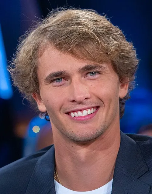 Alexander Zverev'in yakın çekim bir fotoğrafında, dalgalı sarı saçları, parlak mavi gözleri ve lacivert ceketinin üzerinde parlayan altın zinciriyle, koyu mavi bir arka plan önünde bembeyaz dişlerini göstererek içten bir gülümseme sergileyen genç adam.