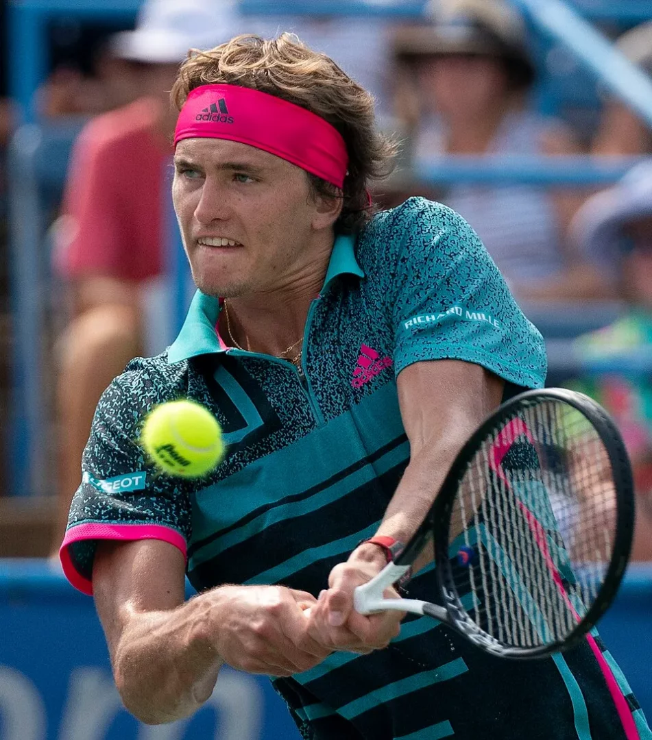 Parlak pembe Adidas saç bandı ve dinamik turkuaz desenli tenis tişörtüyle, gözleri sarı tenis topuna odaklanmış, kortta raketini çift elle kullanarak güçlü bir vuruş yaparken görünen, profesyonel Alman tenisçi Alexander Zverev.