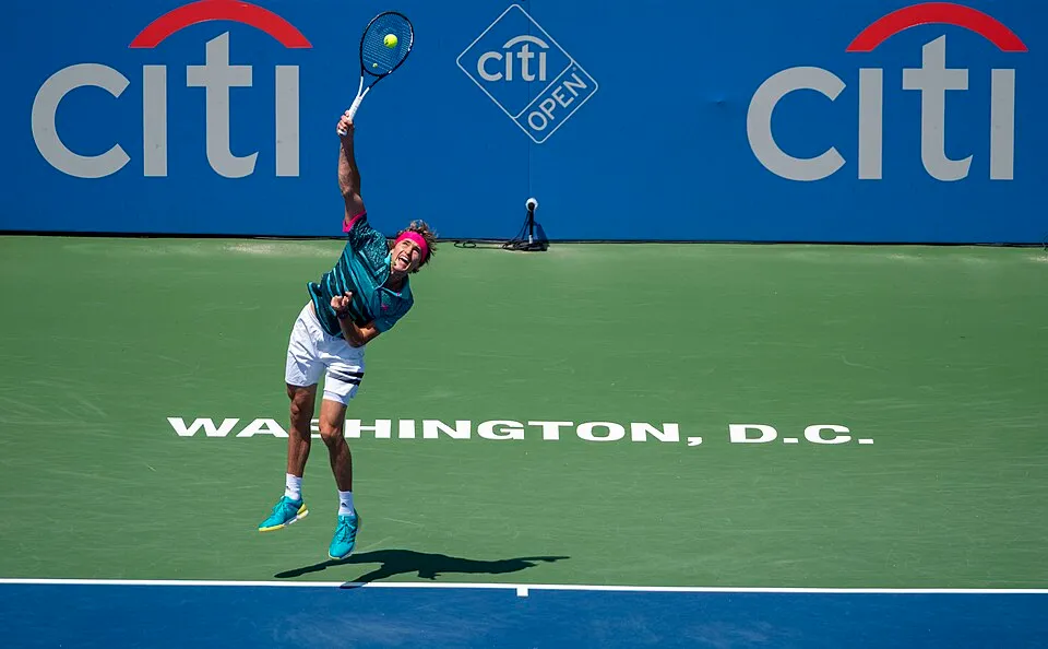 Alexander Zverev, Washington D.C.'deki Citi Open tenis turnuvasında yeşil ve mavi kortta havada yükselerek raketiyle sarı topa güçlü bir servis atışı yapıyor; üzerinde desenli turkuaz bir tişört, beyaz şort ve pembe bir saç bandı var.