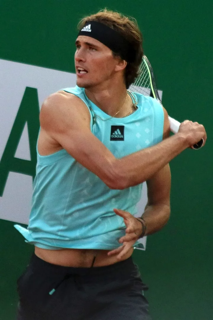 Alexander Zverev, tenis maçında açık mavi kolsuz tişörtü ve siyah Adidas kafa bandı ile tüm gücüyle vuruş yapmaya hazırlanırken, yüzündeki odaklanmış ifadeyle mücadelesini yansıtıyor.