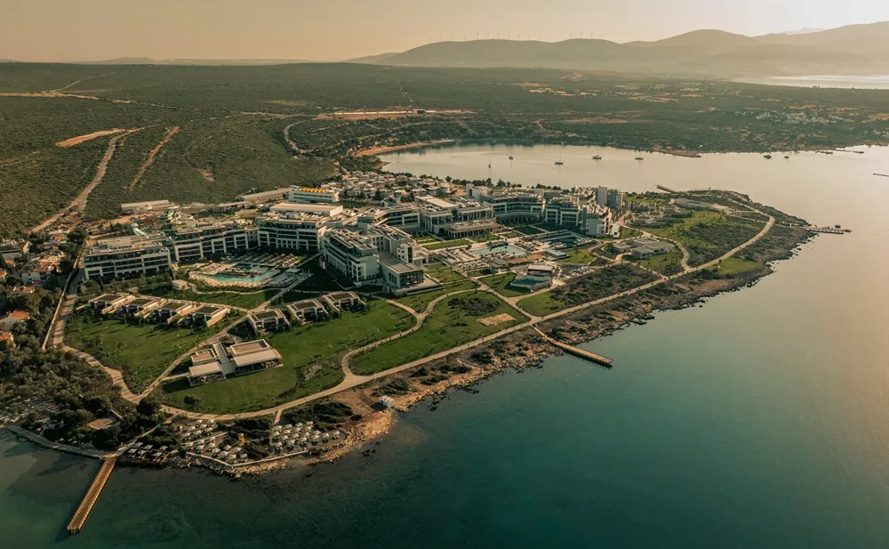 Bir hava fotoğrafı, Ege Denizi'ne uzanan yemyeşil bir yarımada üzerinde konumlanmış, şık ve modern mimarili binalara, açık yüzme havuzlarına, düzenli peyzajlı yeşil alanlara ve iskelelere sahip olan geniş bir tatil köyü olan Anda Barut Collection Didim'i kuşbakışı gösteriyor. Arka planda tepeler ve rüzgar türbinleri görünüyor.