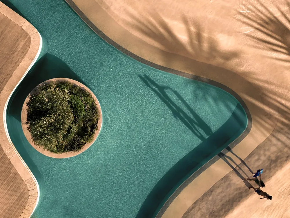 Anda Barut Collection Didim'deki bu kuşbakışı görüntüsünde, turkuaz rengi kıvrımlı yüzme havuzunun ortasında yemyeşil bir ada bulunurken, uzun gölgeler eşliğinde iki kişi havuz kenarında yürüyor.