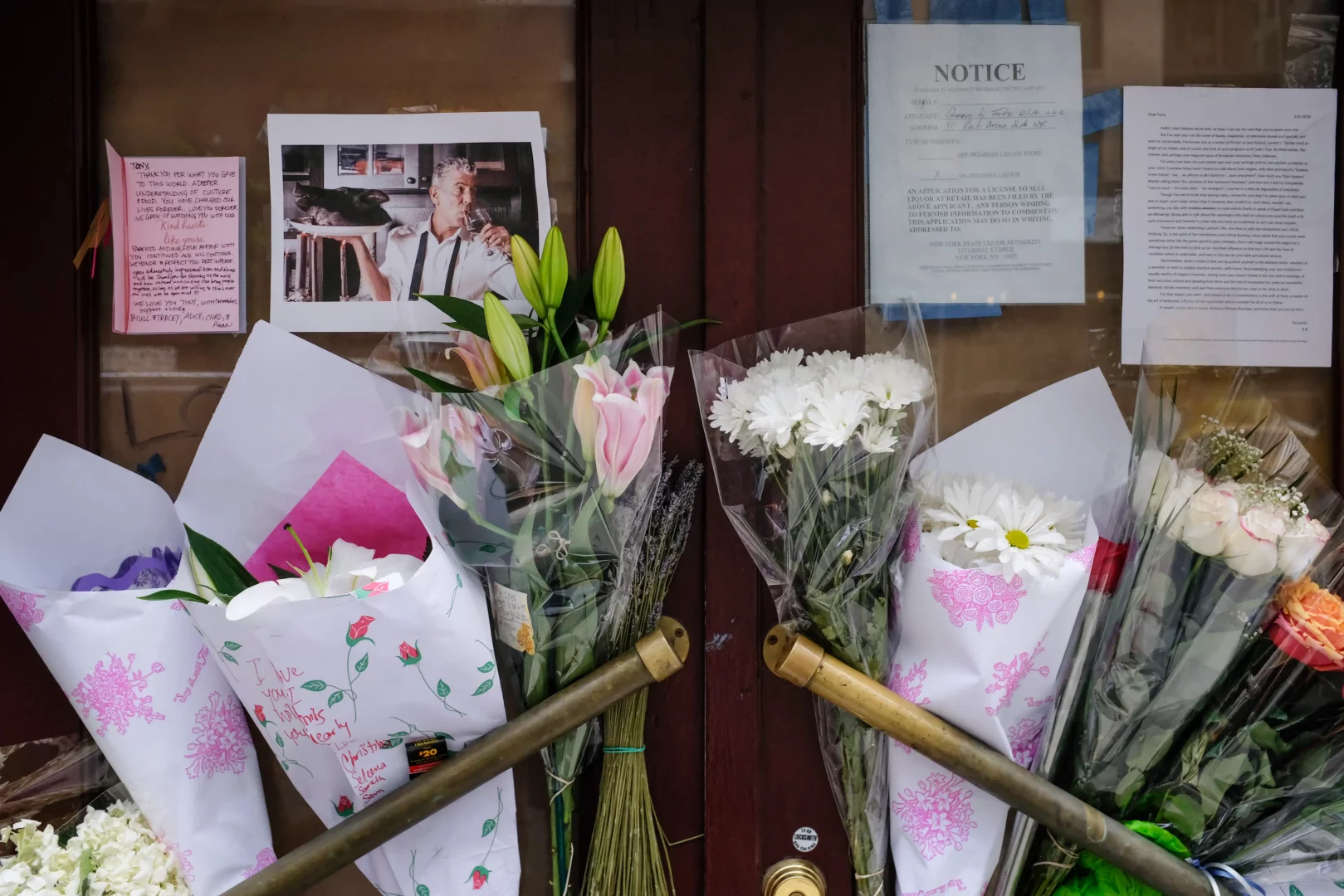 Kapanmış Brasserie Les Halles'in kahverengi kapılarında, Anthony Bourdain'in bir fotoğrafı, üzerinde hayran notları ve beyaz zambak, pembe krizantem ve güllerden oluşan çok sayıda çiçek buketi, onun gastronomi dünyasına yaptığı unutulmaz katkıyı ve modern kaşif ruhunu anmak için toplanmış bir anma yeri görüntüsü sunuyor.