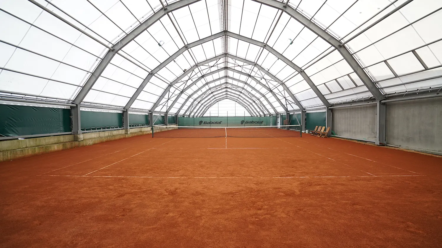 Kemerli ve yarı saydam tavan yapısıyla aydınlık bir kapalı alanda yer alan, ortasında filesi bulunan toprak kort, yani antuka tenis kortu, çevresindeki yeşil bariyerlerde Babolat logoları ve sağda duran birkaç ahşap sandalye ile birlikte modern bir tenis kulübü tesisini gözler önüne seriyor.