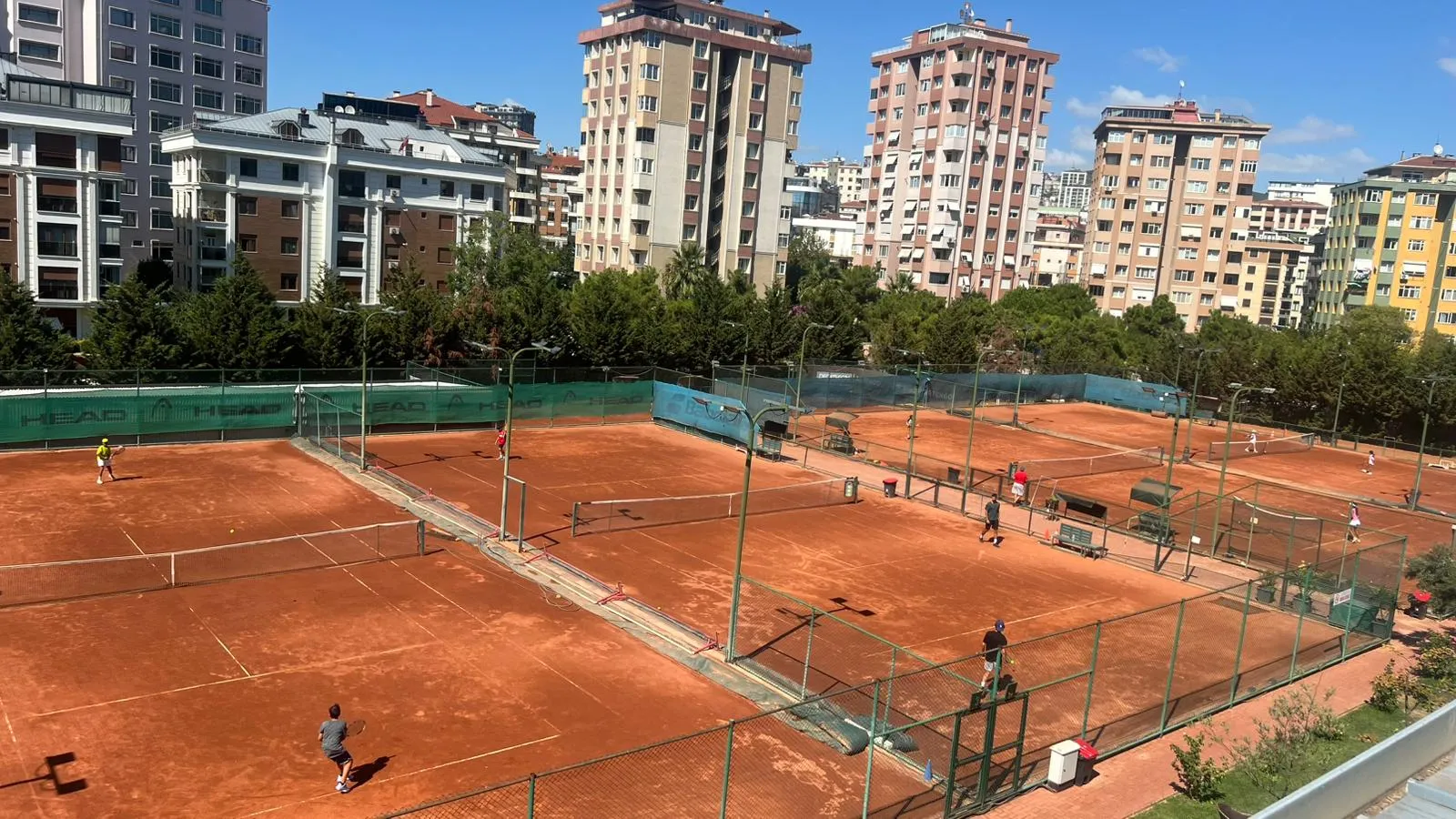 Antuka Tenis Kulübü'nde, şehir silüeti ve yemyeşil ağaçlarla çevrili açık hava tesisinde, güneşli bir günde birçok tenisçinin kırmızı kil kortlarda raketleriyle hareketli bir şekilde tenis oynadığı geniş bir panoramik görünüm.