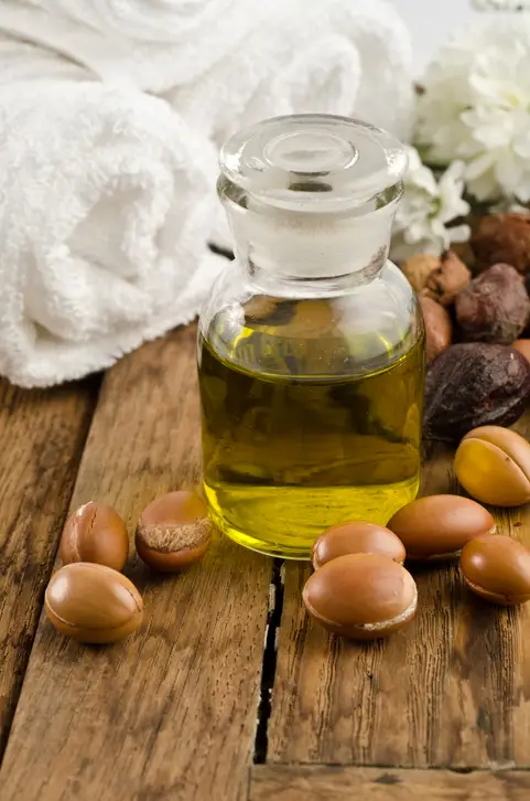 Rustik ahşap bir yüzey üzerinde, içerisine sarımsı-yeşil argan saç yağı doldurulmuş cam bir şişe, etrafına dağınık argan çekirdekleri, beyaz bir havlu ve arka planda narin beyaz çiçeklerle zengin saç bakım yağları sunuyor