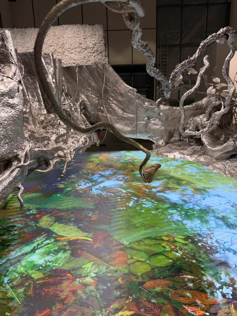Art Basel koleksiyonu içerisinde yer alan bu çağdaş sanat enstalasyonunda, gümüş renkli, kıvrımlı, kök benzeri heykeller, yeşil yapraklar ve sonbahar tonlarındaki batık yaprak yansımalarıyla dolu canlı bir su yüzeyi projeksiyonunun ortasından yükseliyor.