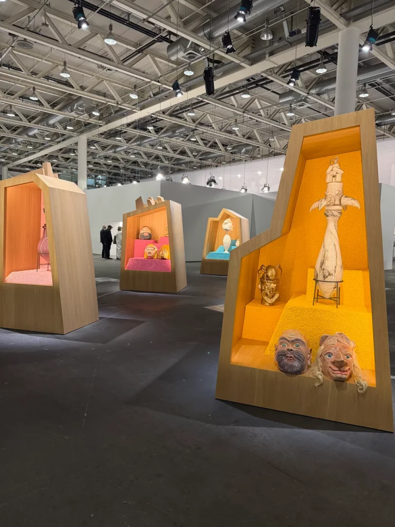 Art Basel’in geniş bir sergi salonunda, sarı, pembe ve mavi tonlardaki zeminlere sahip modern ahşap vitrinlerde, aralarında detaylı maskeler ve stilize heykellerin de bulunduğu çeşitli özgün sanat eserlerinden oluşan bir koleksiyon sergileniyor.