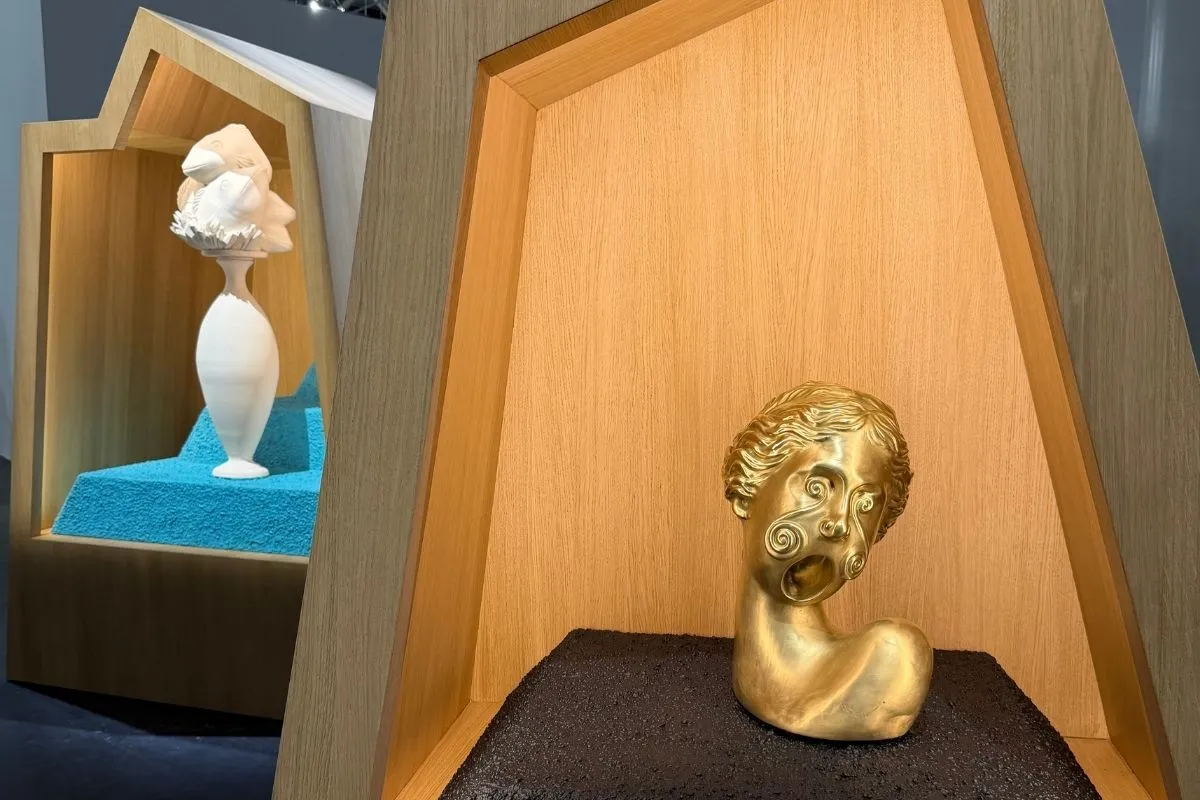 Ahşap nişlerde sergilenen iki modern sanat eseri: sağda, kara dokulu kaide üzerinde duran, ağzı açık, yüzünde spiral desenler olan altın renkli bir insan başı heykeli ve solda, turkuaz renkli geometrik bir zeminde, üst üste binmiş yüzlerden oluşan beyaz vazo benzeri bir heykel, Art Basel koleksiyonundan çarpıcı bir sergilemeyi gösteriyor.