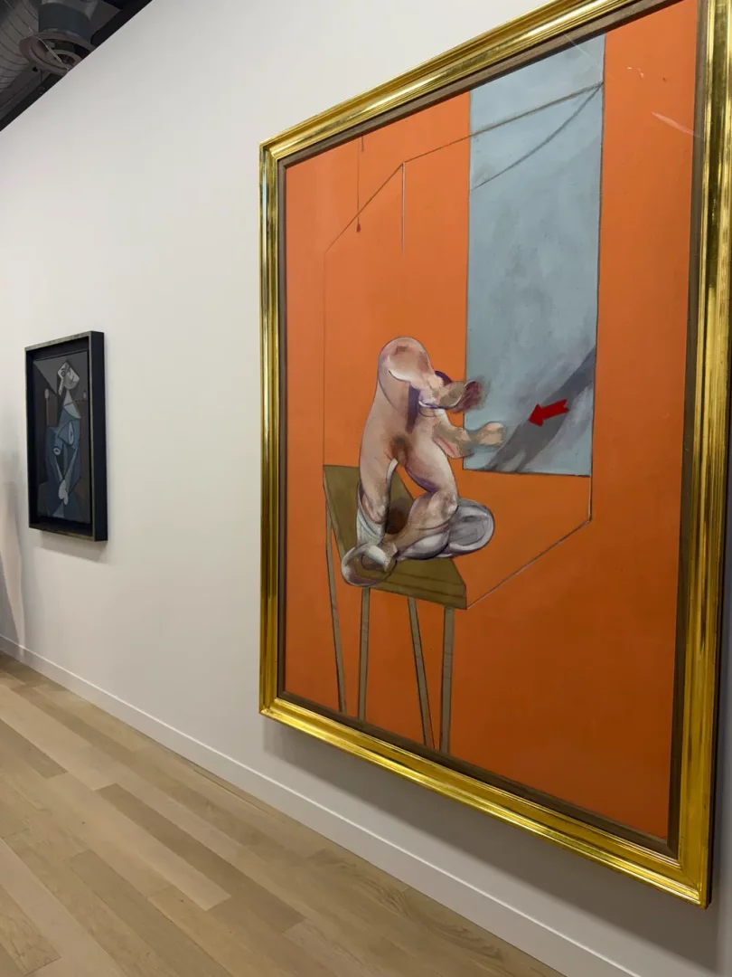 Büyük, altın çerçeveli bir Francis Bacon tablosu, Art Basel koleksiyonu için sergilenen galerinin beyaz duvarında, iskemlede oturan deforme olmuş figürü, canlı turuncu arka planı ve sağdaki mavi bir bölüm ile içindeki kırmızı okuyla dikkat çekiyor; sol arkada ise küçük, koyu çerçeveli soyut bir resim daha asılı duruyor.