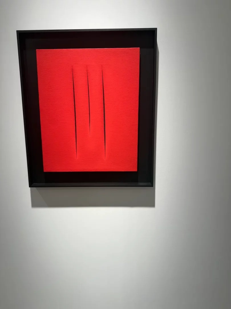 Art Basel koleksiyonundan bir parça olarak sergilenen, parlak kırmızı renkli kare bir tuvalde, tuvalin yüzeyini yaran üç dikey kesiğin bulunduğu, derinlik ve gölge oluşturan minimalist bir sanat eseri sade siyah çerçevesiyle beyaz duvarda asılı duruyor.